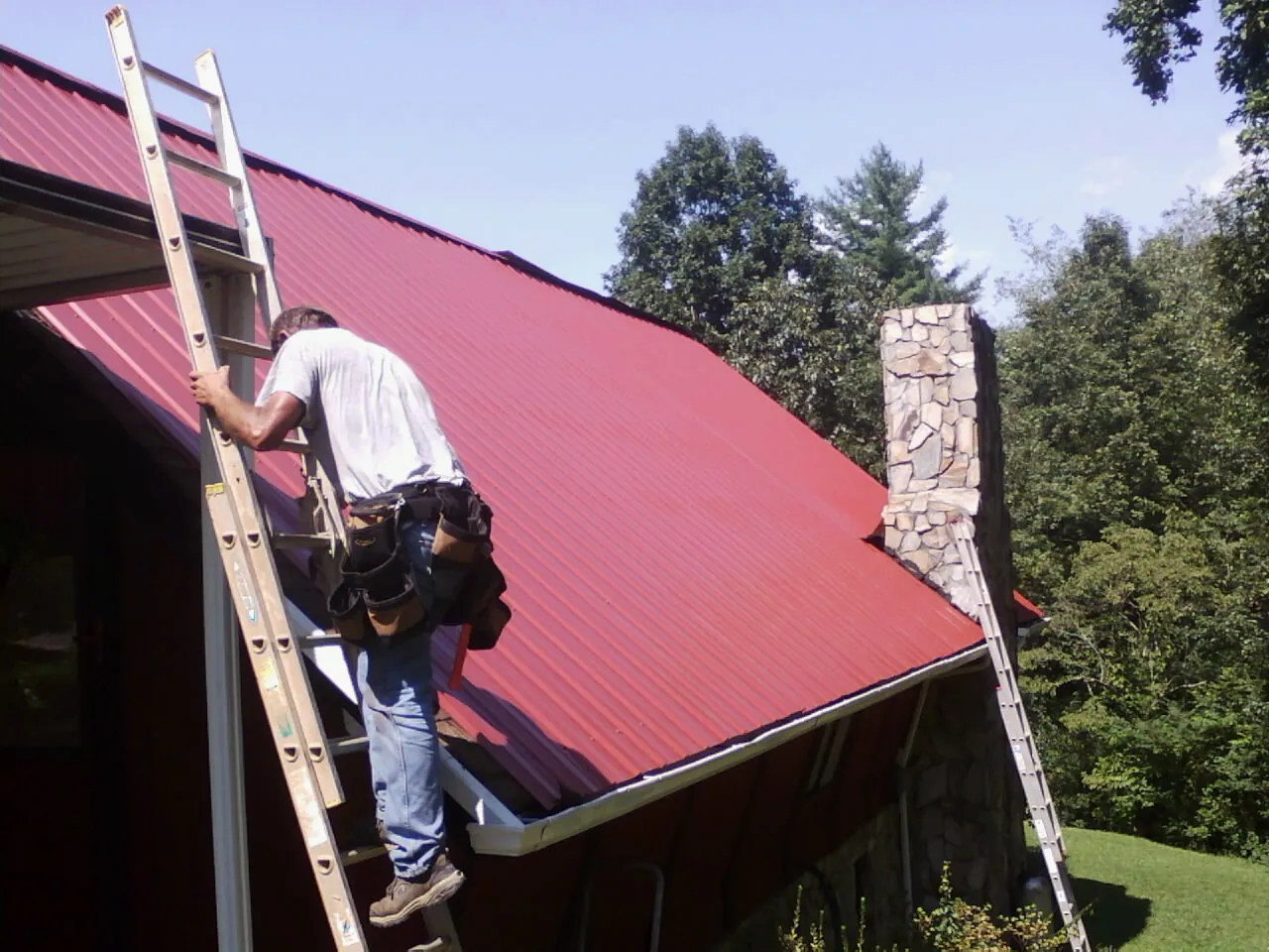 Roofer on a ladder beside a red metal roof and stone chimney — Soffit & Fascia Repair by Roofing Brothers in Nowata